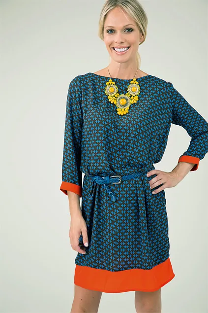 Woman in a blue dress with red accents, wearing a yellow necklace, smiling with hands on hip against a light background.