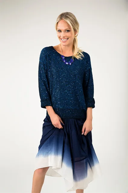 Blonde woman wearing a blue sweater and gradient blue skirt, smiling and standing against a neutral background.
