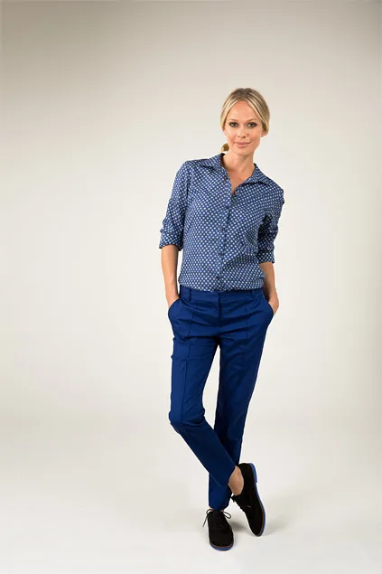 A woman in a blue patterned shirt and matching pants poses with hands in pockets against a neutral background.