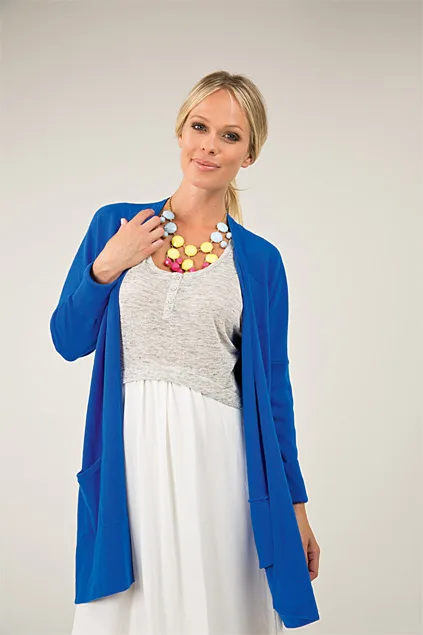 Woman wearing a blue cardigan, gray top, white skirt, and colorful necklace, posed against a plain background.