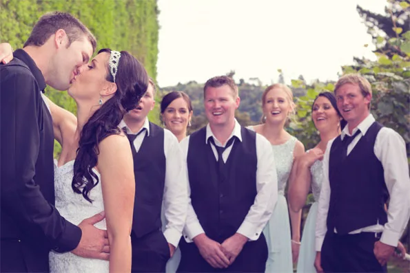Bride and groom kissing with happy bridal party in the background, outdoors.
