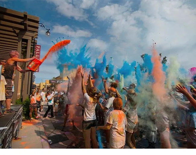 Crowd celebrating with colorful powder thrown in the air at a festival under a partly cloudy sky.
