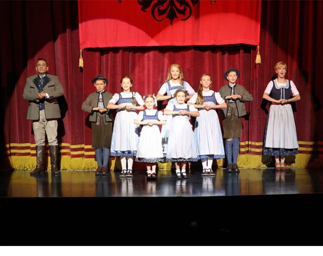 Children in traditional costumes performing on stage, under a red banner with a black emblem.