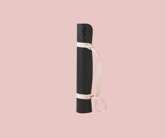 Black rolled yoga mat with a beige carrying strap on a pink background.