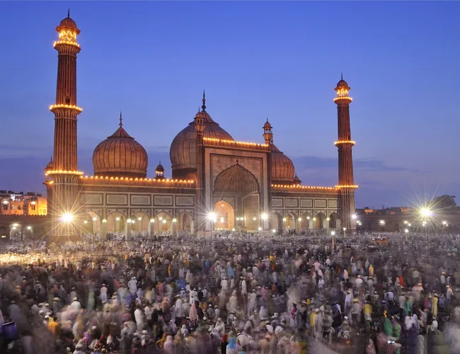 Crowd gathered at Jama Masjid, Delhi, at dusk with the mosque illuminated and clear sky in the background.