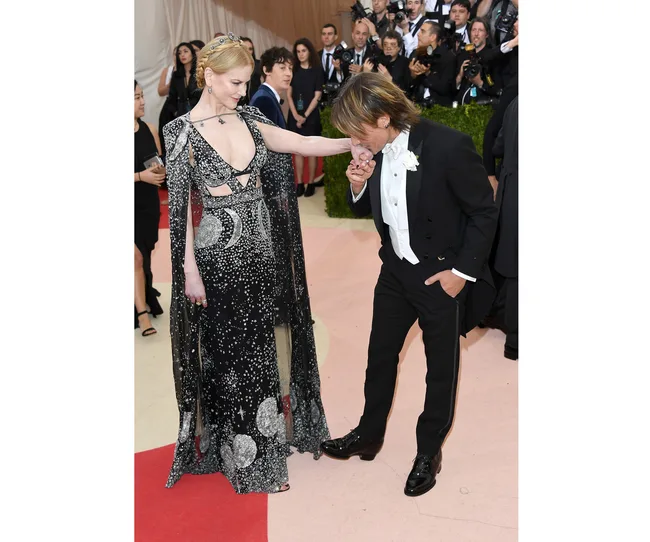 A woman in a starry black gown stands while a man in a tuxedo kisses her hand at a formal event. Photographers are in the background.