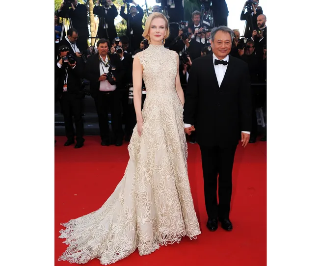 Two individuals posing on a red carpet, with one wearing a white floral gown and the other in a black tuxedo.