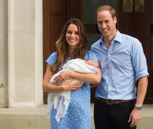 A couple smiling, with the woman holding a newborn wrapped in a white blanket, standing outside a building.