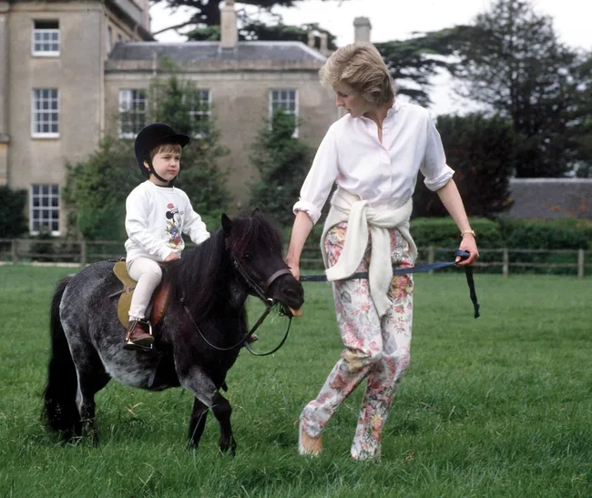 Woman leads a small pony on which a child wearing a helmet is sitting, set in a grassy field near a large building.