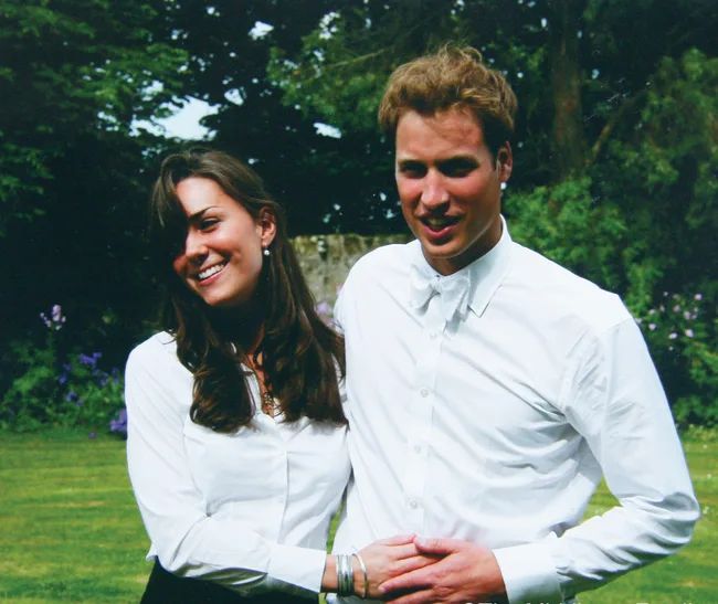 A smiling couple in white shirts stands outdoors, surrounded by greenery.
