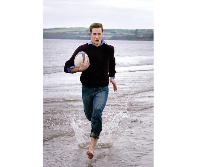 A person running barefoot through the water on a beach, holding a rugby ball with a calm sea in the background.