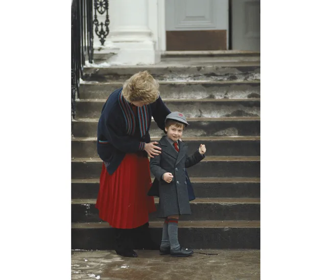 A woman holds a young boy's shoulder as they stand on outdoor steps; the boy is in a gray coat and cap.