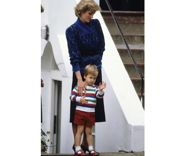 A woman in a blue sweater stands with a child in a striped sweater and red shorts waving by white stairs.