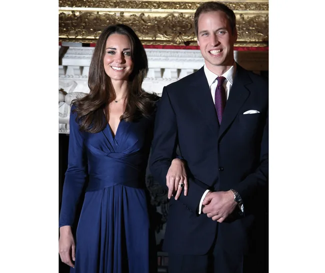 A woman in a blue dress and a man in a suit stand arm-in-arm, smiling, against an ornate background.