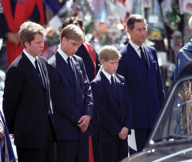 A group of four men in black suits stand solemnly at an outdoor event, with flowers visible in the background.