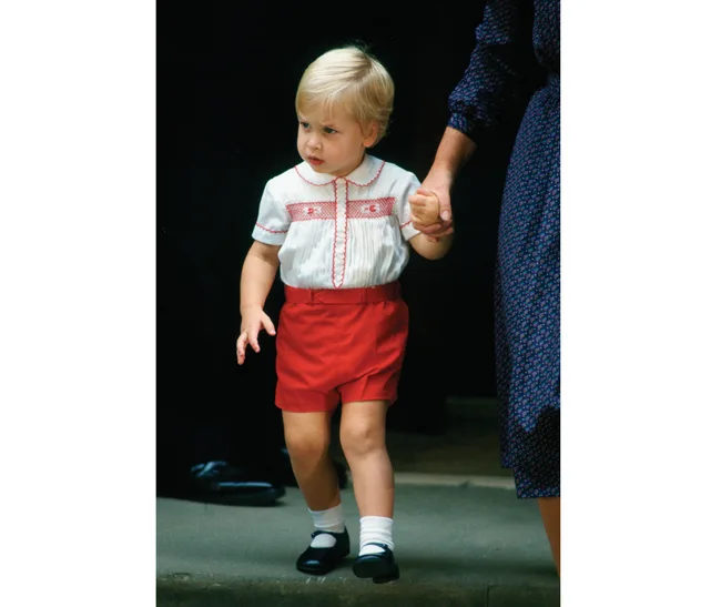 Toddler in red shorts and white shirt, holding an adult's hand, walking on a step.