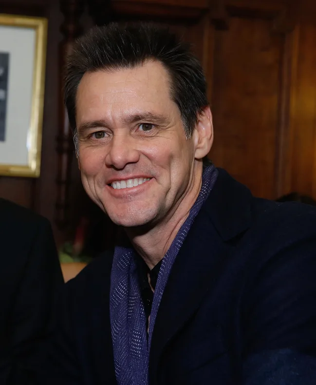 A person smiling in a warmly lit room, wearing a dark jacket and scarf.