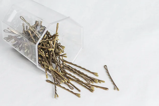 Plastic container of bronze bobby pins, some spilled on white surface.