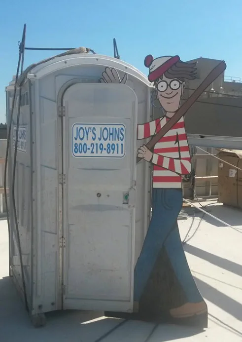 Cartoon character cutout next to a porta-potty labeled "Joy's Johns" with a contact number, on a sunny rooftop.