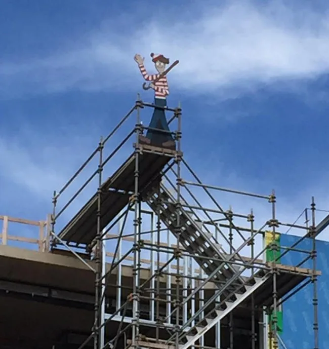Cartoon character in striped shirt and beanie on scaffolding against blue sky.