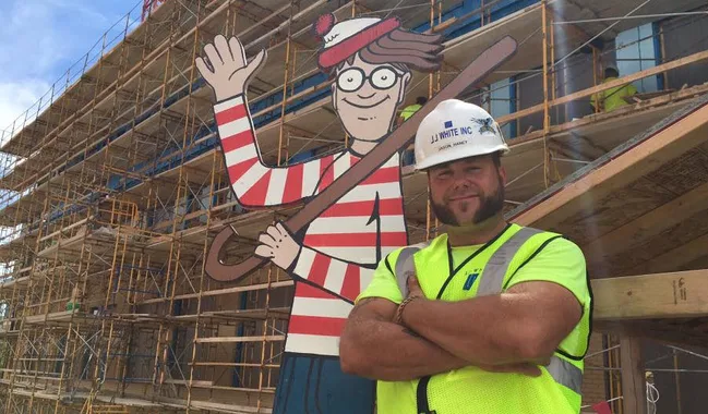 Construction worker posing with large "Where's Waldo?" figure in front of scaffolding.