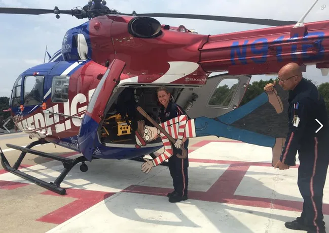 Medical crew with cardboard cutout of "Where's Waldo?" next to red and blue medical helicopter on helipad.