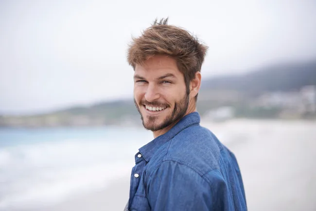 Smiling man with short hair and a beard wearing a denim shirt on a beach.