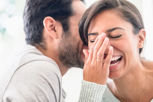 A man whispers in a woman's ear, making her laugh while covering her mouth with her hand.