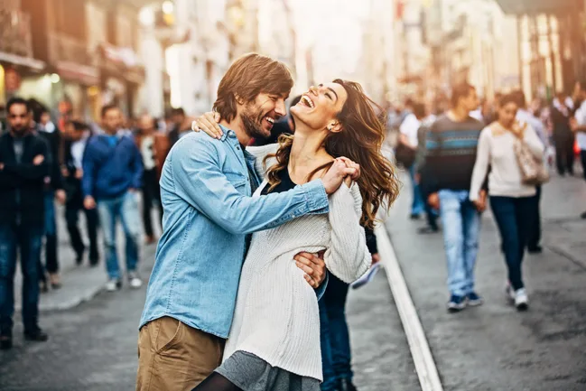 Couple laughing and dancing on a busy city street, surrounded by a bustling crowd.