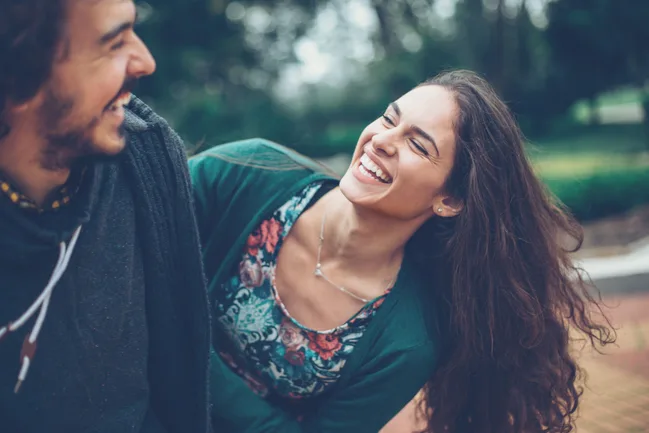 A woman with long hair and a floral dress laughs with a man outdoors.