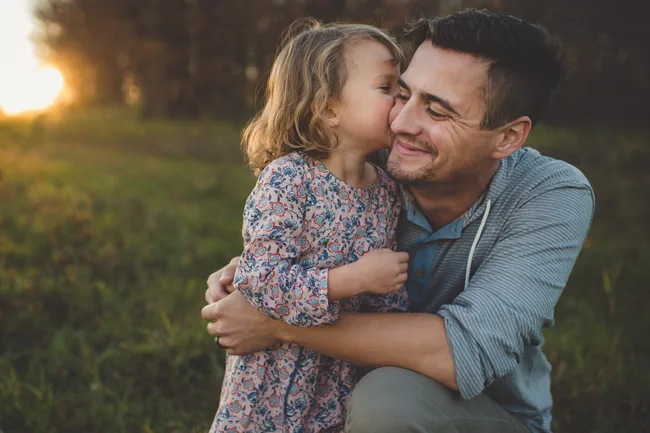 Father hugging daughter in floral dress, smiling at sunset in a field.