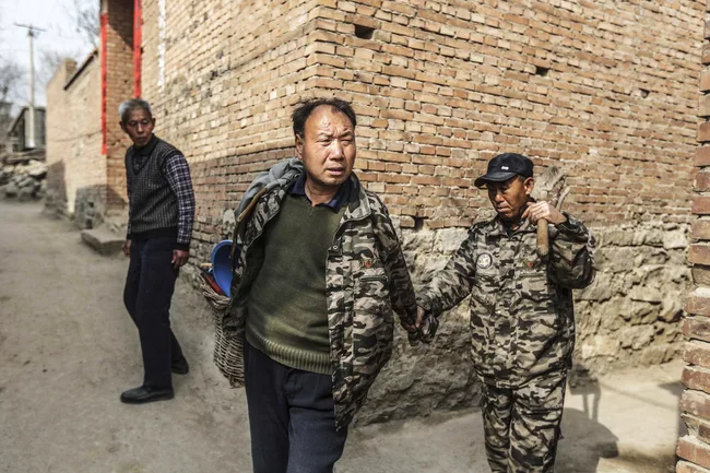 Two men in camouflage jackets walk along a dirt path near a brick wall; another man stands nearby watching them.