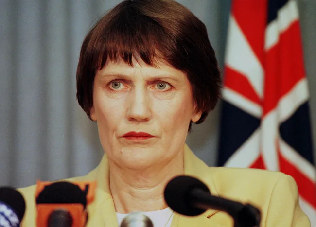 Portrait of a woman speaking at a press conference with microphones and a flag in the background.