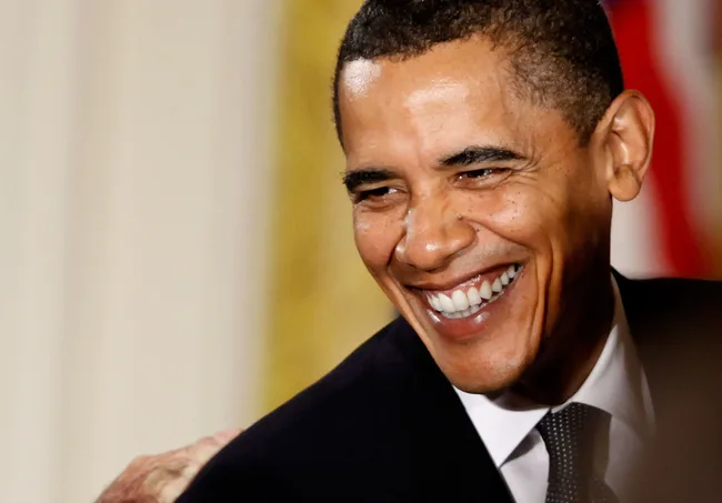 A man in a suit smiling broadly, set against a blurred background.