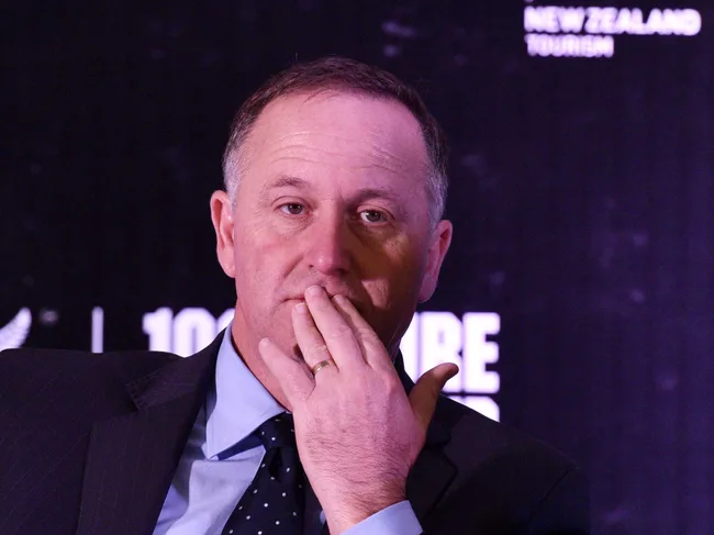 A person in a suit resting their hand on their face, with a New Zealand tourism banner in the background.