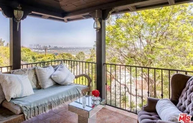 Covered terrace with a cozy sofa, pillows, a glass-top table, and a view of trees and a distant city skyline.