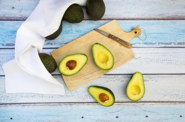 Avocados halved on a wooden board with a knife, surrounded by whole avocados on a rustic blue table.