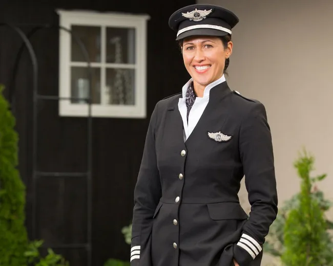 Female pilot in a uniform smiling, with a building and greenery in the background.