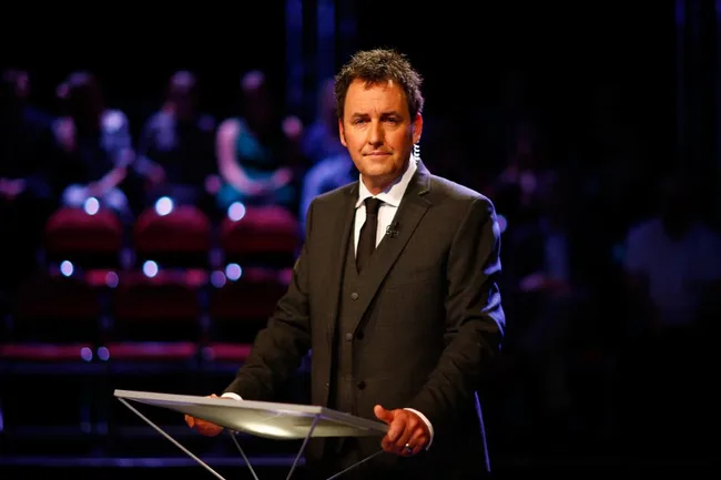 A man in a dark suit stands at a podium on a dimly lit stage with an audience in the background.