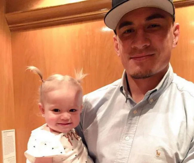 Man in a cap holding a smiling toddler with pigtails in front of a wooden wall.