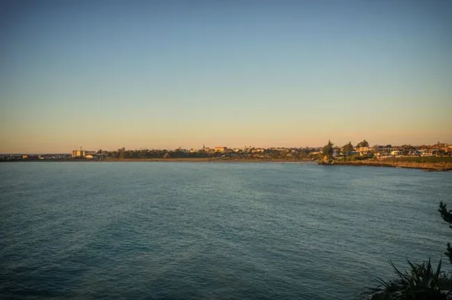 Calm ocean with distant shoreline and buildings under a clear sky at sunset.