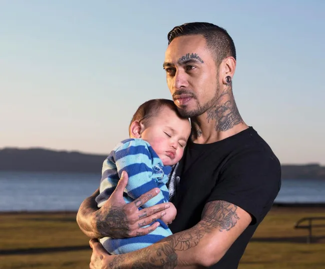 Man with tattoos holding a sleeping baby by the ocean.