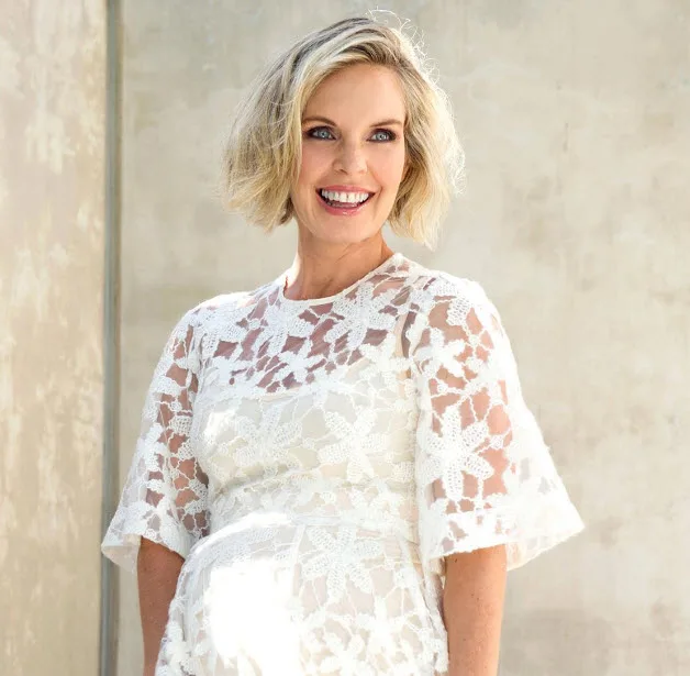 A smiling woman in a white lace dress, standing against a plain wall.