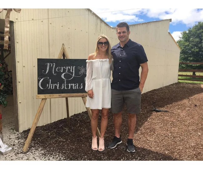 A couple poses beside a "Merry Christmas" sign outdoors on a sunny day.