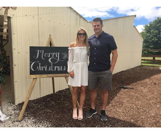 A couple poses beside a "Merry Christmas" sign outdoors on a sunny day.