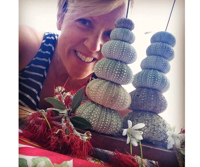 A person smiling next to a stack of sea urchin shells with flowers at the base, wearing a striped tank top.
