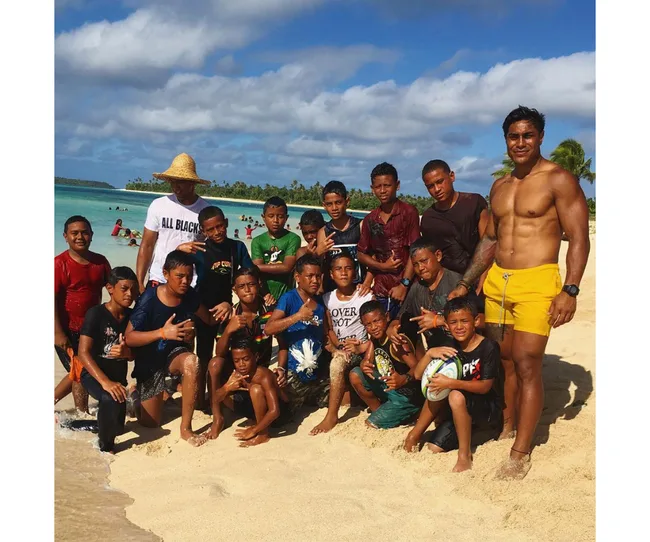 Group of people, including kids with a rugby ball, on a sunny beach with turquoise water and palm trees in the background.