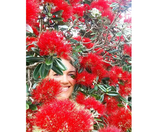 Smiling woman partially hidden among vibrant red pohutukawa flowers and green leaves.