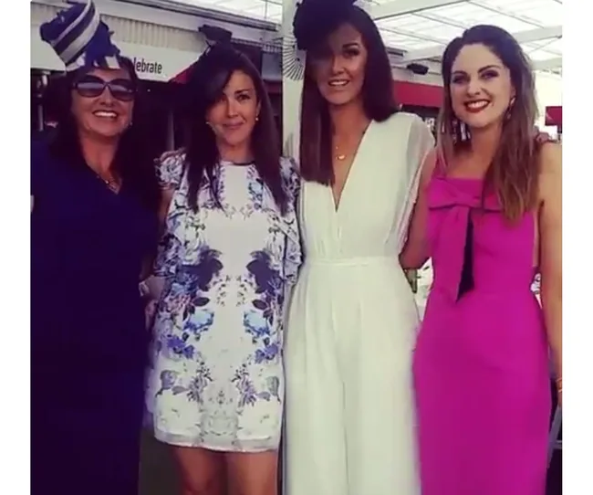 Four women in elegant dresses and hats, smiling and posing together at a social event.
