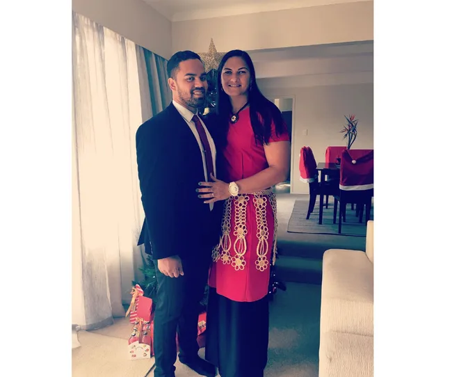 A couple dressed formally stands together in a living room adorned with Christmas decorations.